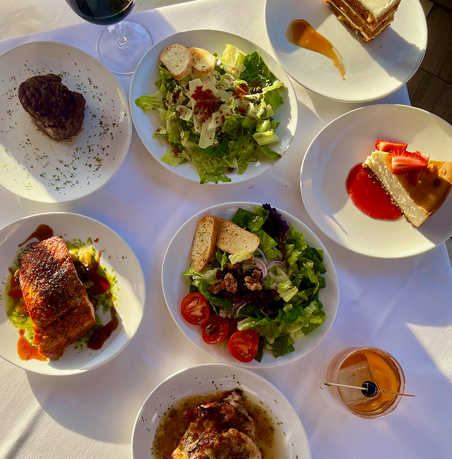 An overhead view of an elegant dinner spread on a white tablecloth featuring multiple plated dishes including a seared salmon fillet, filet mignon steak, Caesar salad with croutons, a walnut and mixed greens salad with cherry tomatoes, chicken in brown sauce, a slice of strawberry cheesecake, a layered tiramisu cake, and craft cocktails with garnishes, fine dining restaurant table setting, warm ambient lighting.