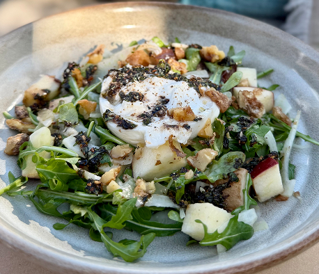 An overhead shot of a gourmet arugula salad with burrata cheese, roasted walnuts, sliced pears, balsamic reduction, and microgreens on a rustic ceramic plate, warm restaurant table setting with soft natural lighting.
