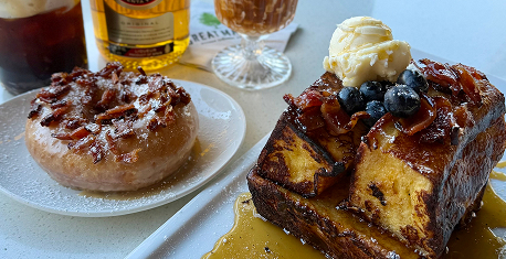 A brunch spread featuring a glazed cinnamon roll with cream cheese frosting and a decadent French toast topped with blueberries, whipped butter and maple syrup, alongside a cold brew coffee, sunny casual restaurant table, warm golden hour lighting.